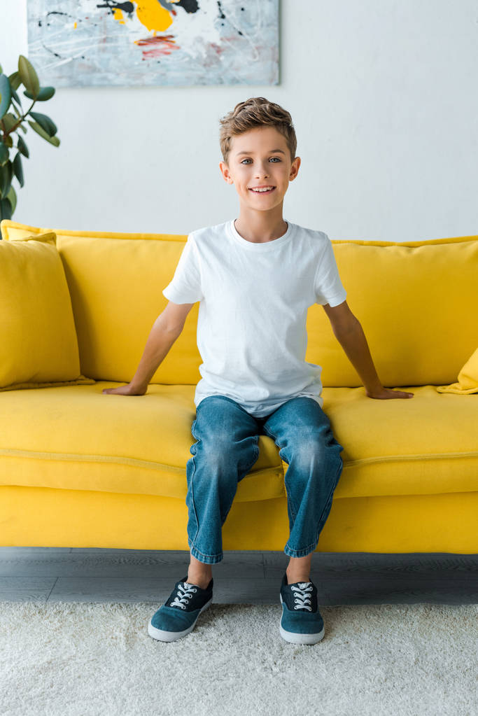 χαρούμενο αγόρι με λευκό t-shirt κάθεται σε κίτρινο καναπέ  - Φωτογραφία, εικόνα