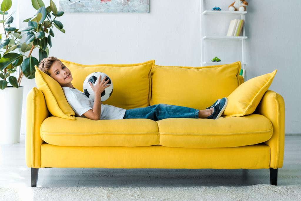 šťastný chlapec v bílém tričku ležící na žluté pohovce s fotbalem  - Fotografie, Obrázek