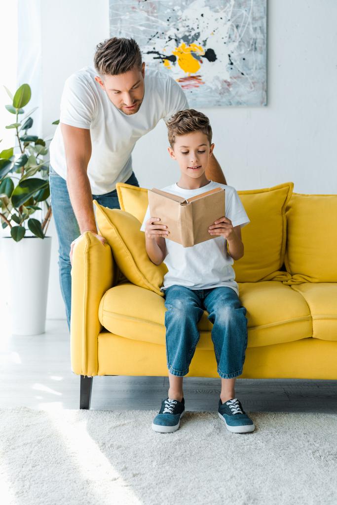 handsome father standing near son reading book while sitting on sofa  - Photo, Image