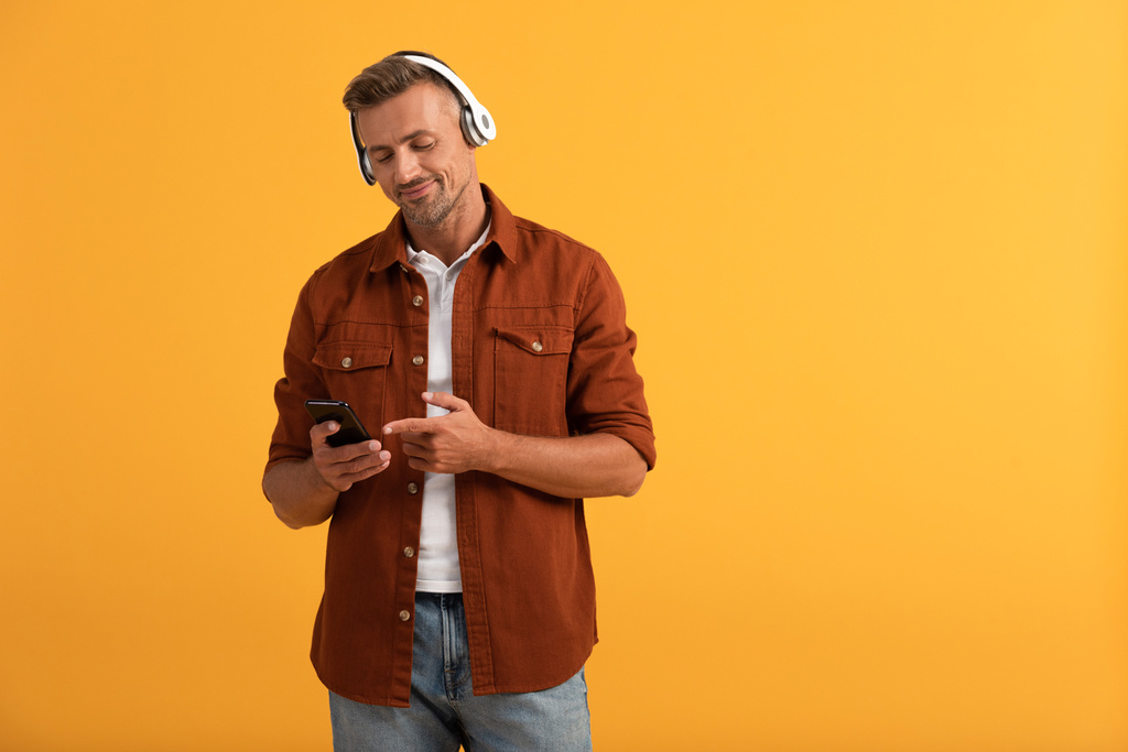 šťastný muž poslouchající hudbu a ukazující prstem na smartphone izolované na oranžové  - Fotografie, Obrázek