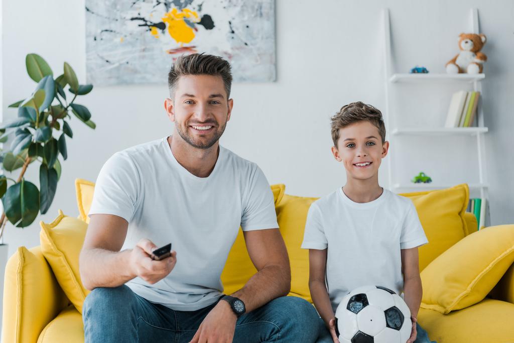 alegre padre sosteniendo mando a distancia cerca de hijo con fútbol sentado en el sofá
  - Foto, Imagen