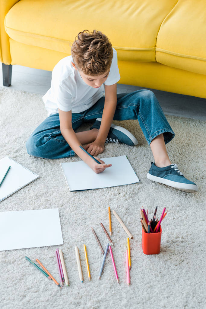 χαριτωμένο παιδί κάθεται στο χαλί κοντά σε κίτρινο καναπέ και σχέδιο σε χαρτί στο σπίτι  - Φωτογραφία, εικόνα