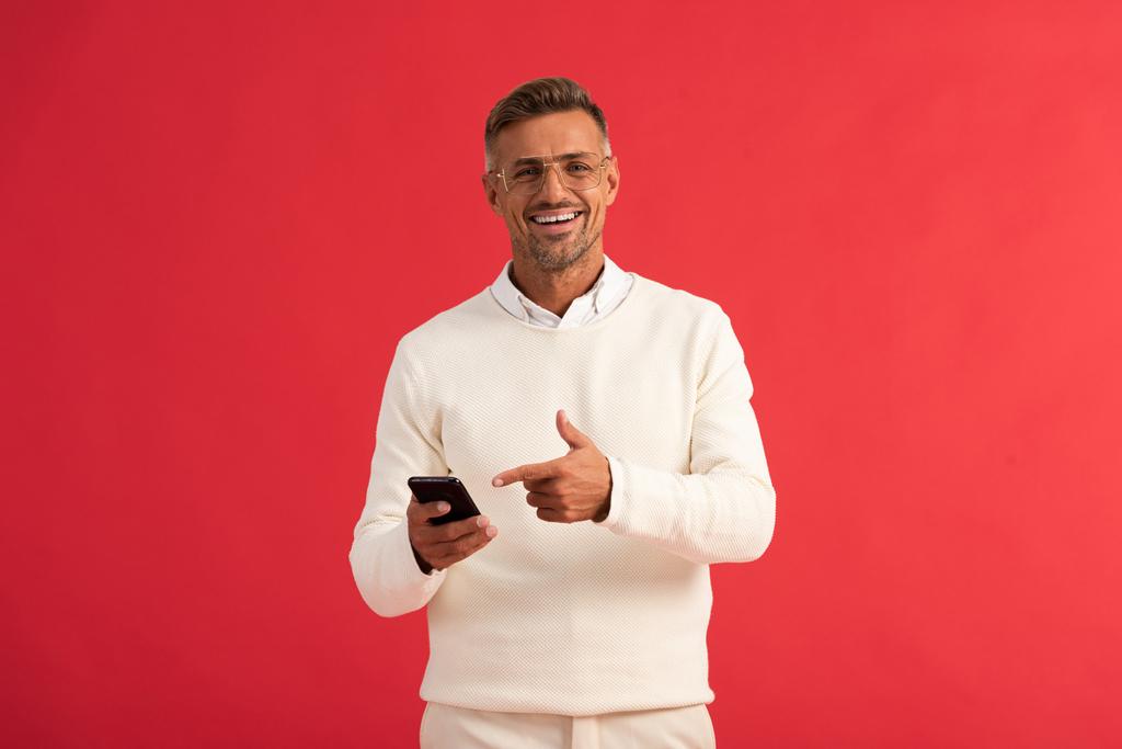 homme heureux dans des lunettes pointant du doigt le smartphone isolé sur rouge
  - Photo, image
