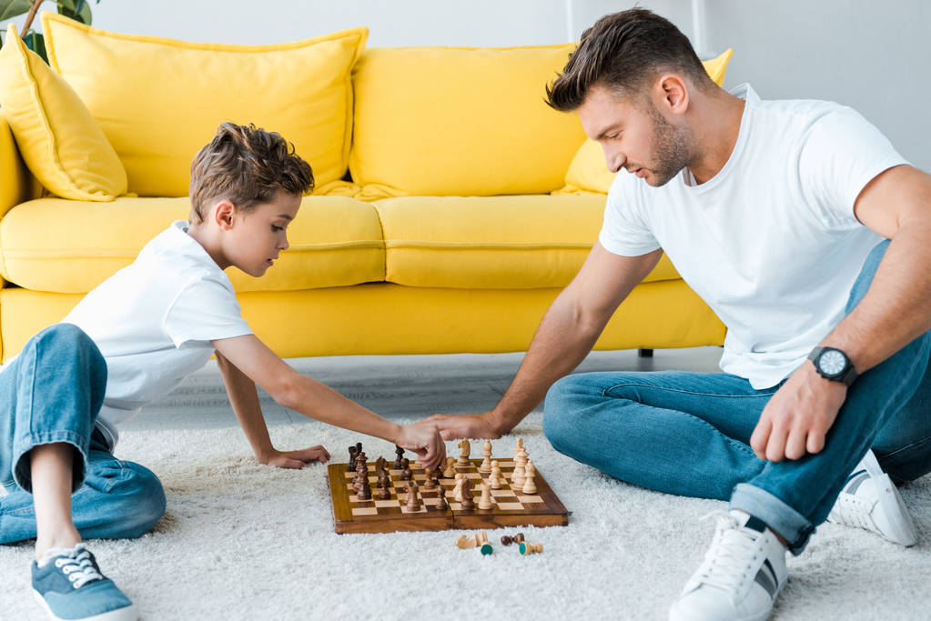 πλαϊνή άποψη του όμορφου πατέρα και του γιου που παίζουν σκάκι στο χαλί  - Φωτογραφία, εικόνα
