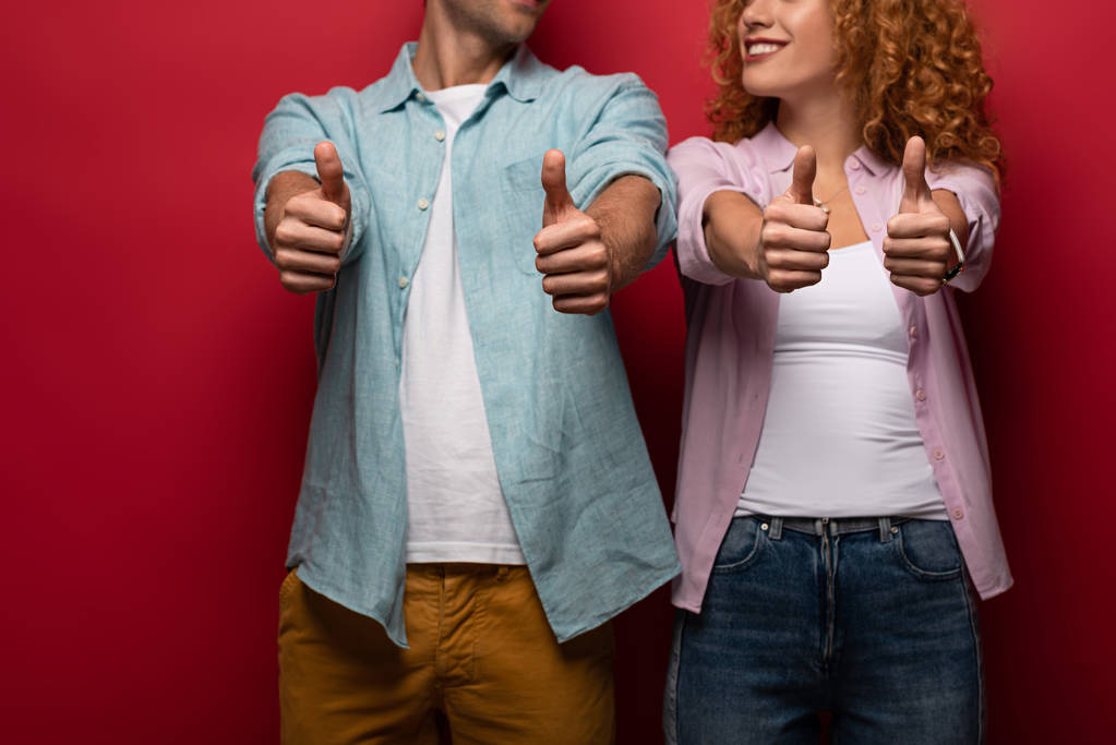 Обрезанный вид улыбающейся пары, показывающей большие пальцы вверх, изолированной на красном
 - Фото, изображение