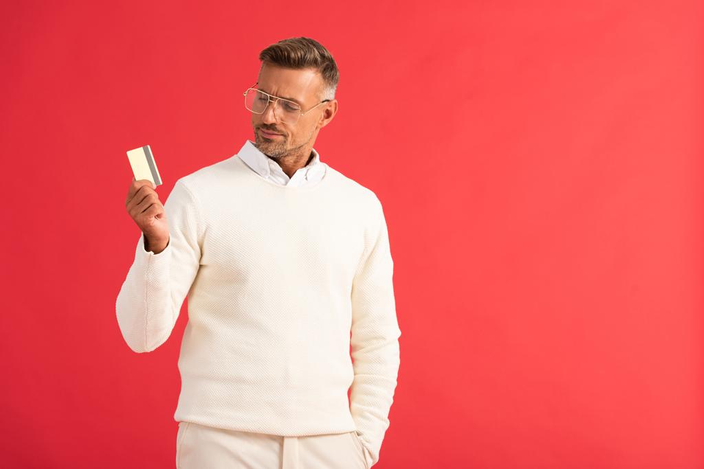 κομψό άνθρωπος σε γυαλιά κρατώντας πιστωτική κάρτα απομονώνονται σε κόκκινο  - Φωτογραφία, εικόνα