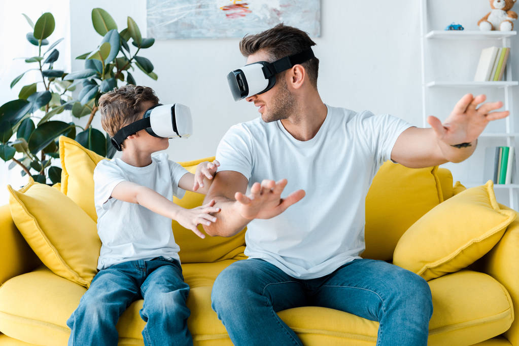 gelukkig vader en zoon in virtual reality headsets gebaren in de woonkamer  - Foto, afbeelding