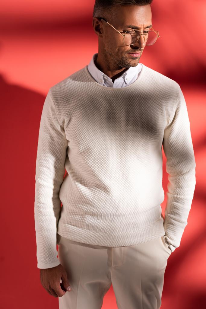 homme élégant dans des lunettes debout avec la main dans la poche sur rouge avec des ombres
  - Photo, image