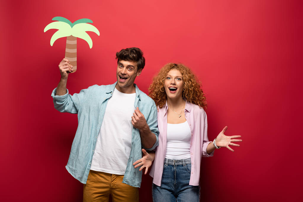 couple of cheerful travelers holding paper palm, isolated on red - Photo, Image