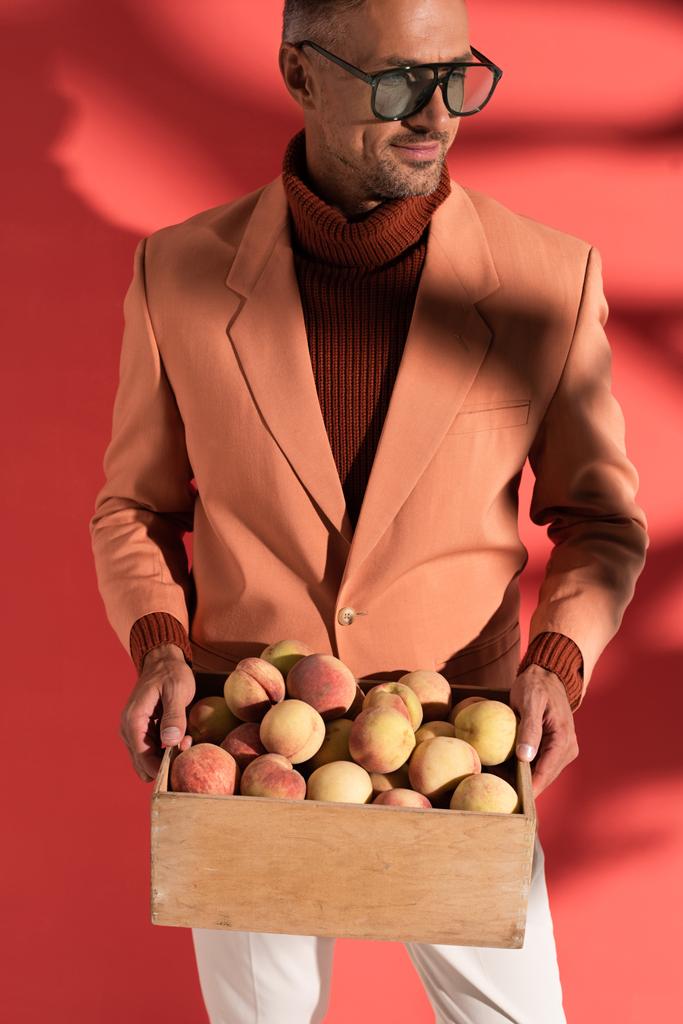 glücklicher Mann in Sonnenbrille mit Schachtel mit süßen Pfirsichen auf Rot mit Schatten - Foto, Bild