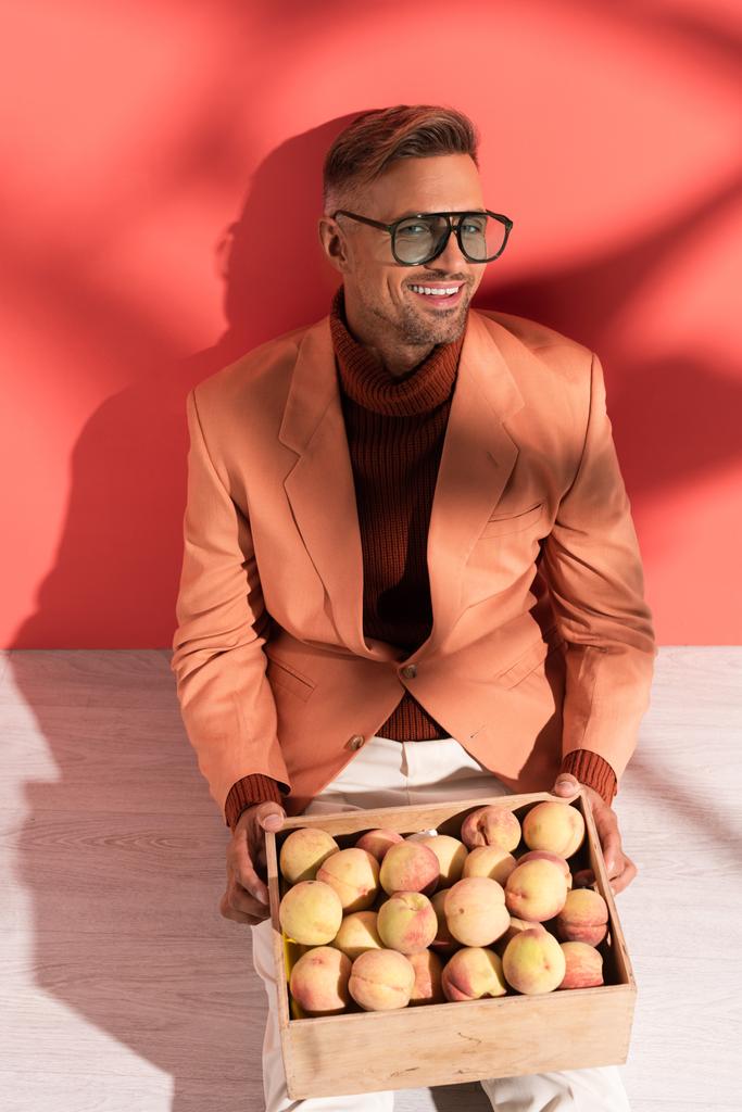 happy man in blazer sitting with sweet peaches in box on red and white with shadows - Photo, Image