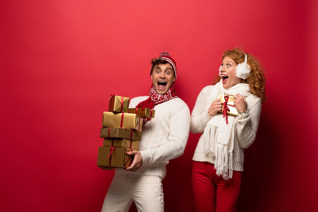 Aufgeregtes Paar im Winteroutfit mit Weihnachtsgeschenken auf Rot - Foto, Bild