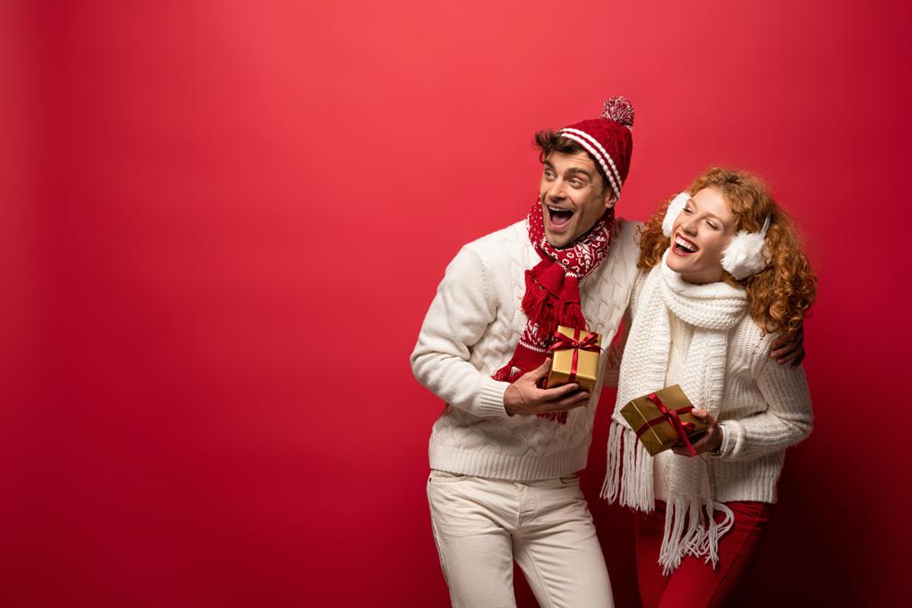 όμορφο ευτυχισμένο ζευγάρι σε χειμωνιάτικο ντύσιμο κρατώντας χριστουγεννιάτικα δώρα που απομονώνονται σε κόκκινο - Φωτογραφία, εικόνα