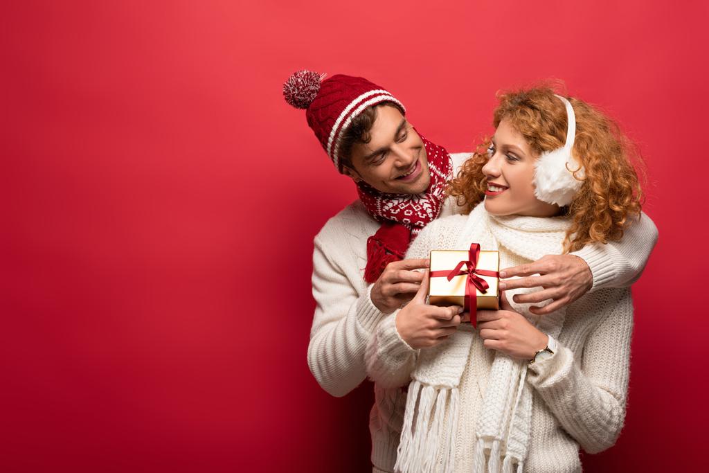 casal feliz em roupa de inverno segurando presente de Natal isolado no vermelho
 - Foto, Imagem