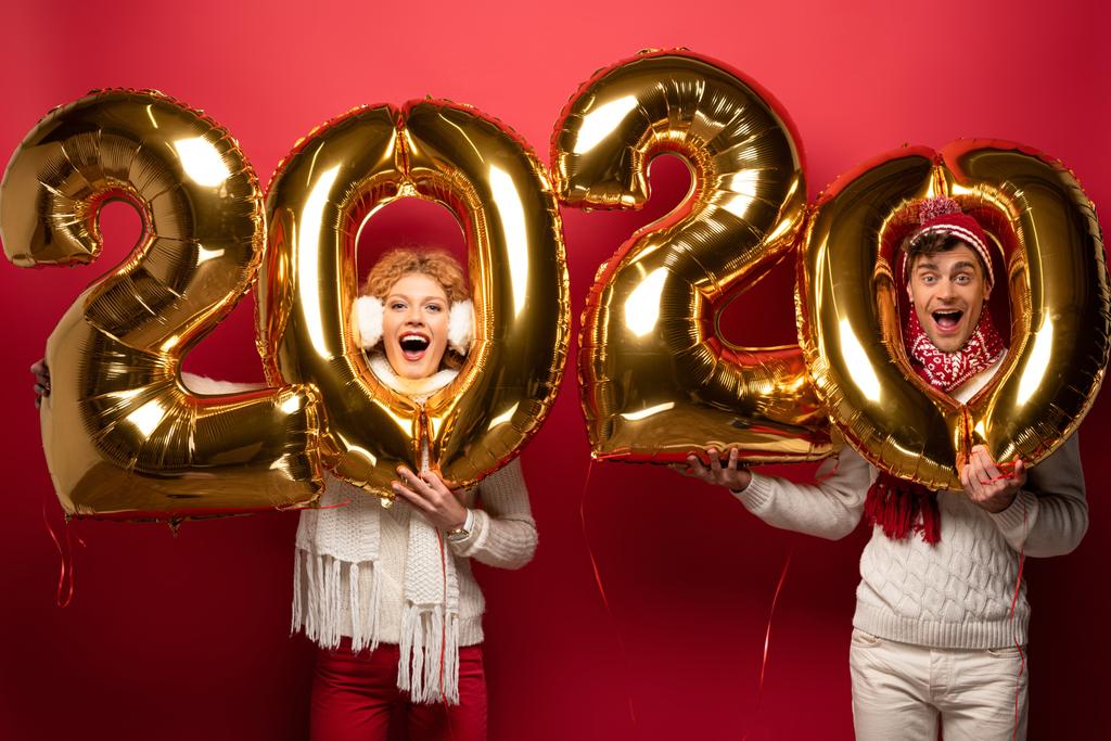 vzrušený pár v zimním oblečení drží nový rok 2020 zlaté balónky, na červené - Fotografie, Obrázek