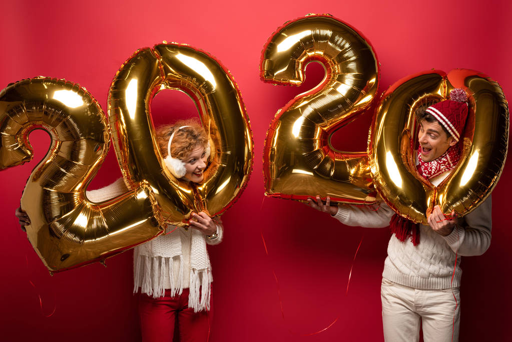 έκπληκτο ζευγάρι σε χειμερινό ντύσιμο εκμετάλλευση νέο έτος 2020 χρυσά μπαλόνια, σε κόκκινο - Φωτογραφία, εικόνα