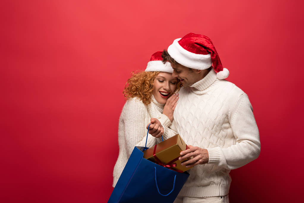 heureux couple dans chapeaux Santa avec présent dans le sac à provisions, isolé sur rouge
 - Photo, image