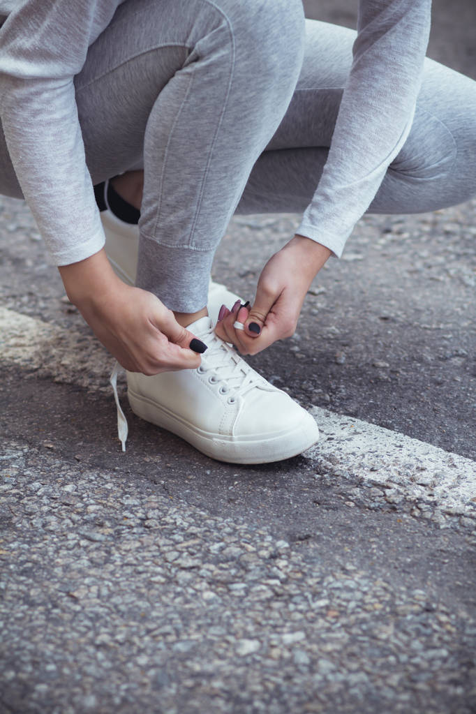 Девушка присела на корточки, чтобы завязать шнурки на белых кроссовках на асфальтовой дороге, осень спортивная концепция на открытом воздухе
 - Фото, изображение