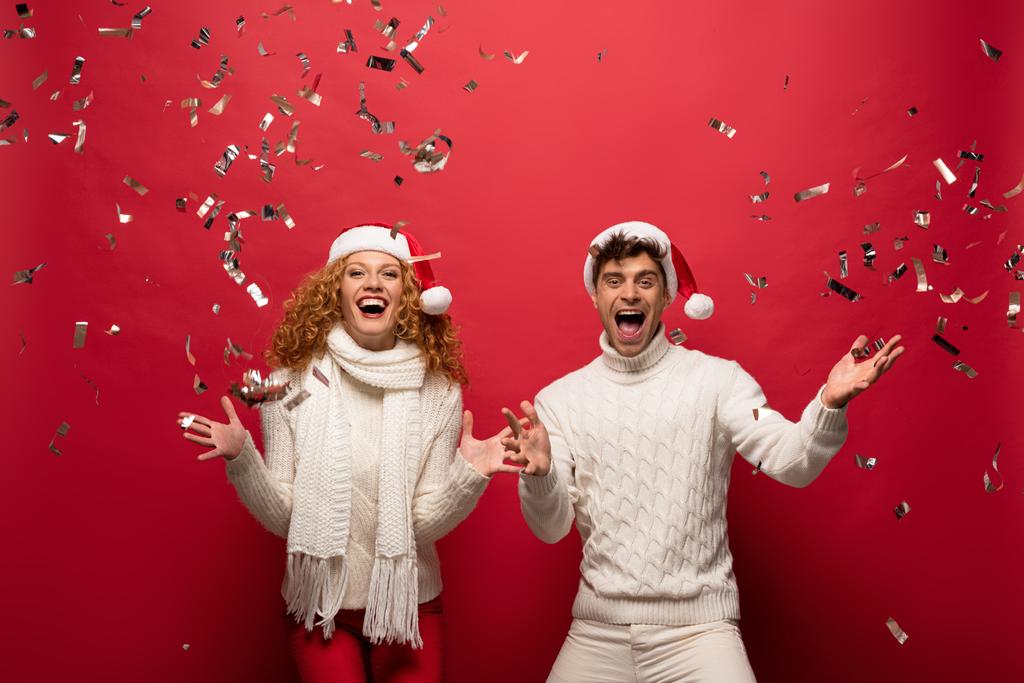 excited couple in santa hats screaming and celebrating with golden confetti, isolated on red - Photo, Image