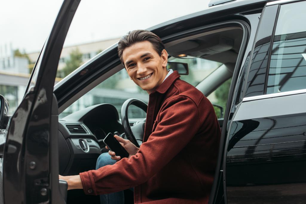 Lächelnder Taxifahrer sitzt im Auto mit offener Tür und hält Smartphone in der Hand - Foto, Bild