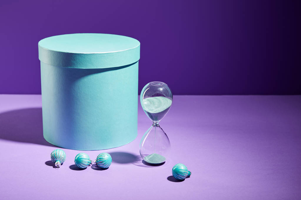 caja de regalo azul y bolas decorativas y reloj de arena sobre fondo púrpura
 - Foto, Imagen