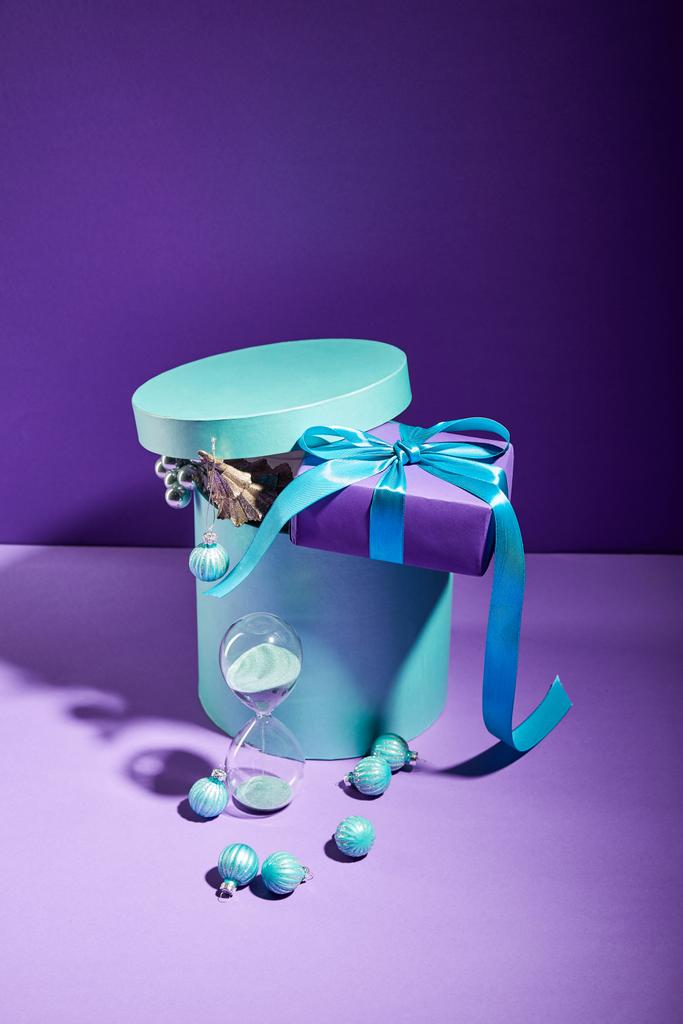 синя коробка Різдвяна прикраса і подарунок біля пісочного годинника на фіолетовому фоні
 - Фото, зображення