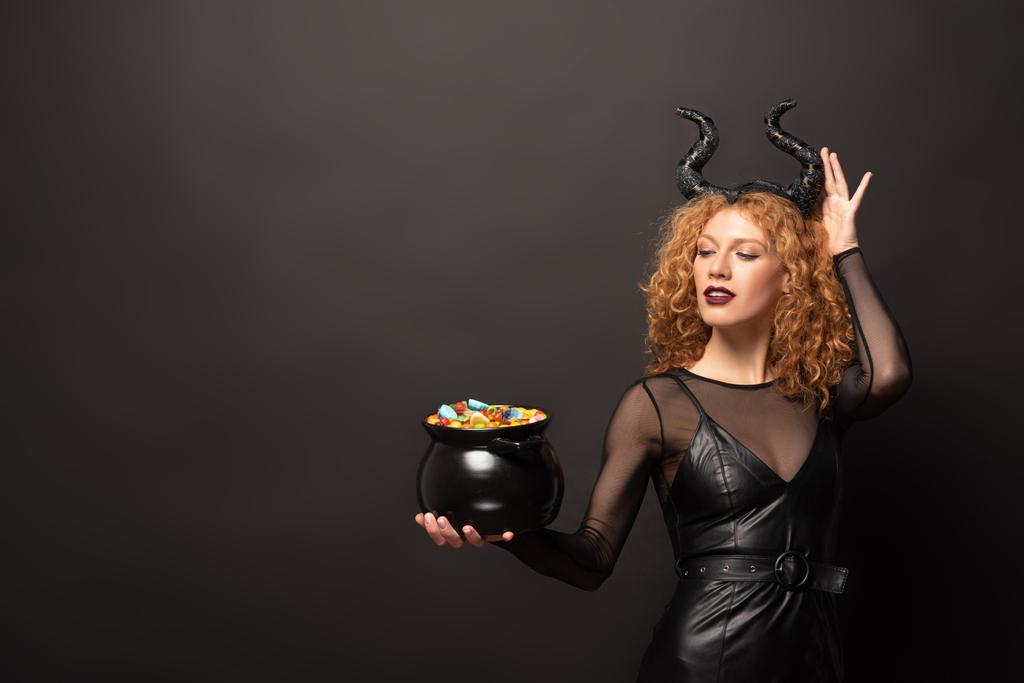 aantrekkelijke vrouw in kwaadaardige kostuum holding pot met snoepjes voor Halloween op zwart - Foto, afbeelding