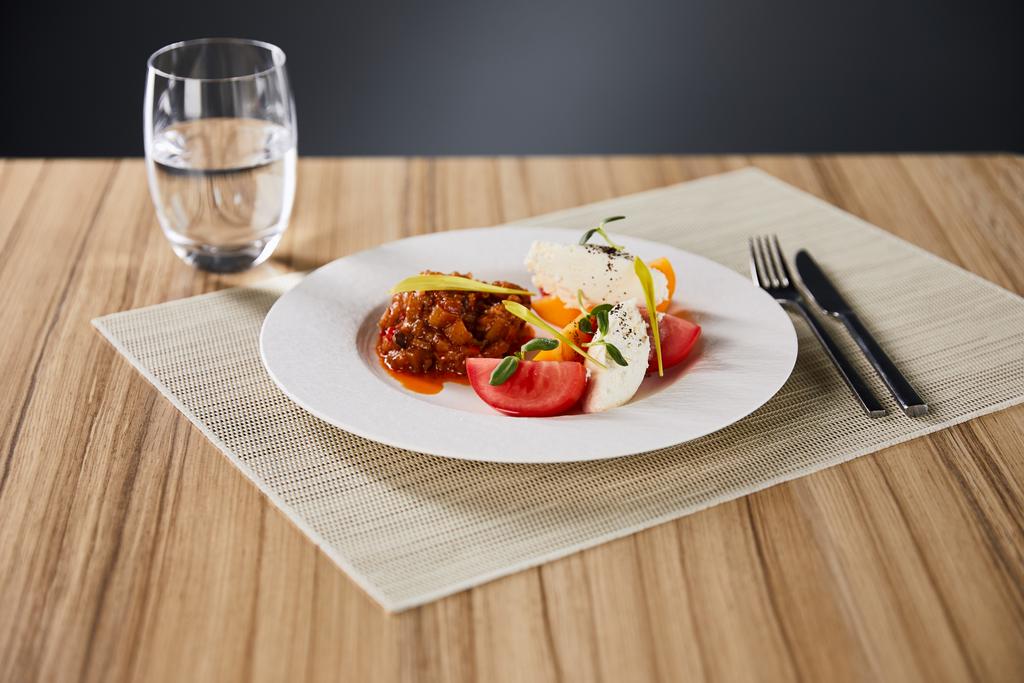 селективный фокус изысканного ресторанного блюда с баклажанной икрой и помидорами, подаваемых на деревянном столе с водой и столовыми приборами, изолированными на сером
 - Фото, изображение