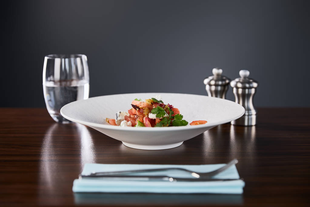 szelektív fókusz ízletes éttermi saláta sajttal szolgált fehér tányéron fa asztal vízzel, evőeszközök és só és bors shakers fekete alapon - Fotó, kép