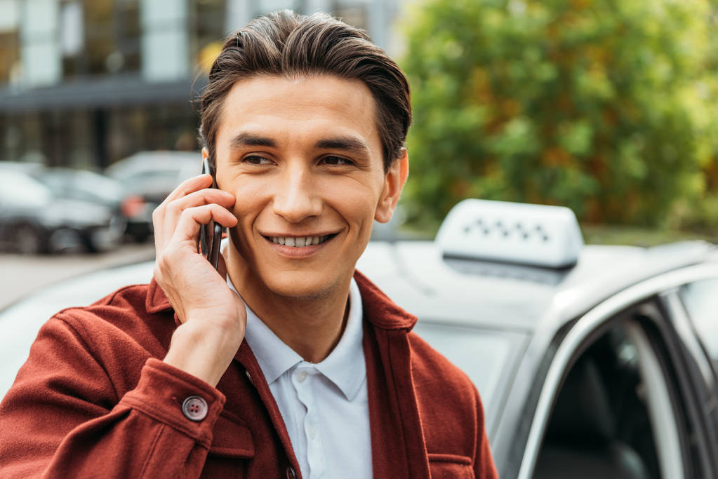 Selektiver Fokus eines lächelnden Mannes, der auf dem Smartphone mit Taxi im Hintergrund spricht - Foto, Bild