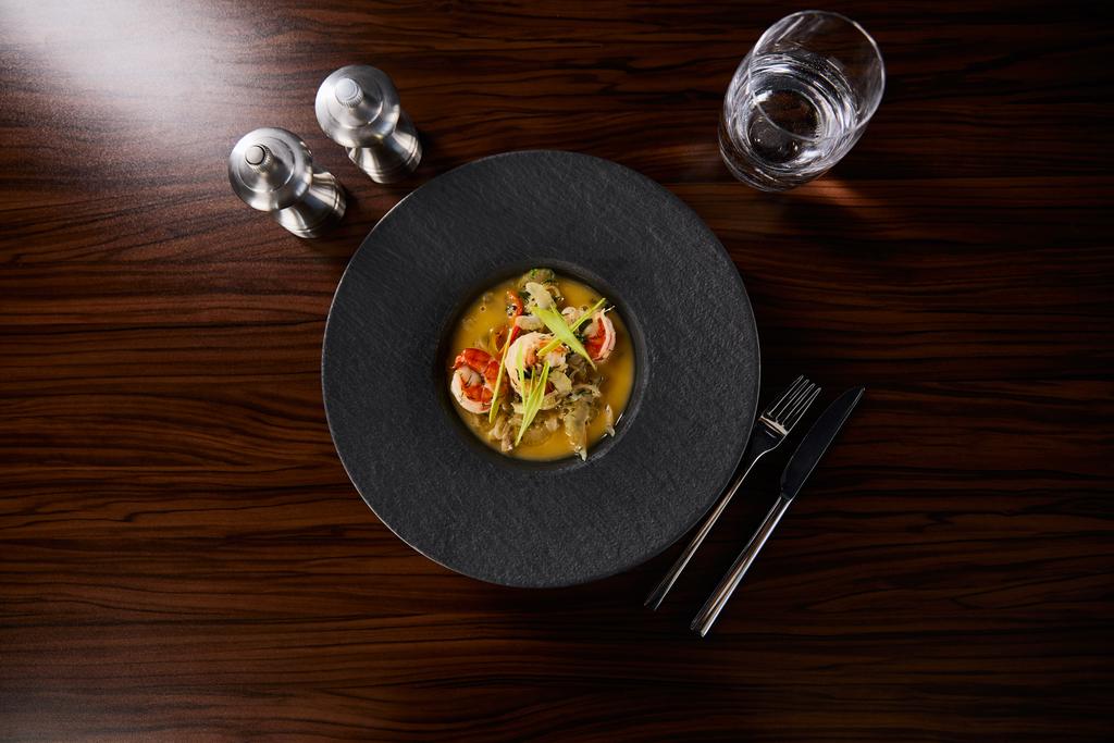 вид зверху смачний ресторанний суп з креветками в чорній тарілці, що подається на серветці з столовими приборами та водою
 - Фото, зображення