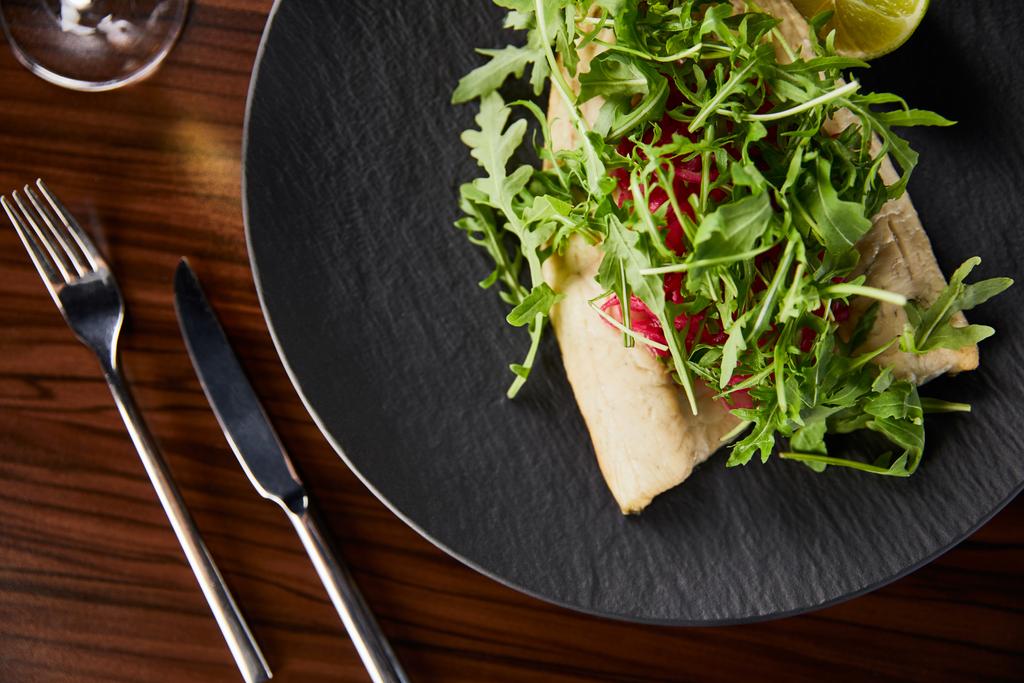 Lezzetli restoran balığı bifteği, kireç ve roka, ahşap masada, çatal bıçak takımının yanında. - Fotoğraf, Görsel
