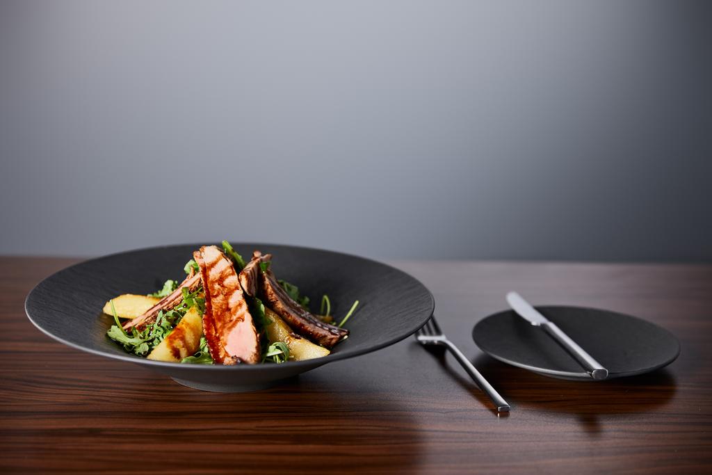 νόστιμη ζεστή σαλάτα με ρόκα, πατάτα και κρέας σε μαύρο πιάτο σε ξύλινο τραπέζι κοντά σε μαχαιροπήρουνα σε γκρι φόντο - Φωτογραφία, εικόνα