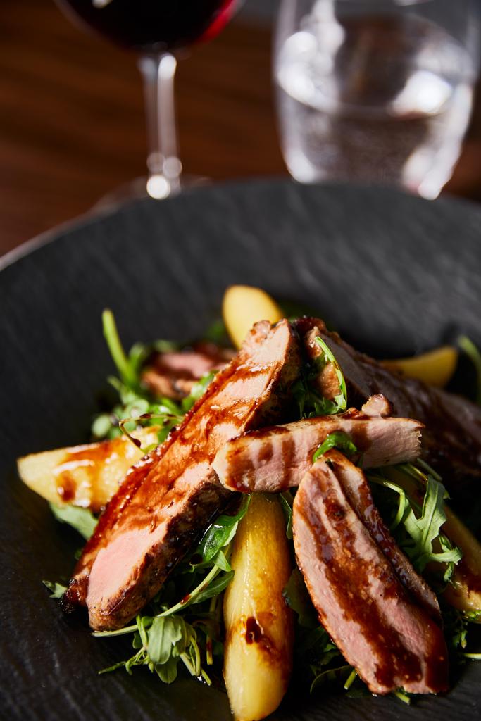 close-up uitzicht op heerlijke warme salade met arugula, aardappel en vlees in zwarte plaat op houten tafel - Foto, afbeelding