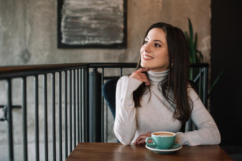 sognante giovane donna sorridente seduta al tavolo di legno con cappuccino sul balcone in caffetteria
 - Foto, immagini