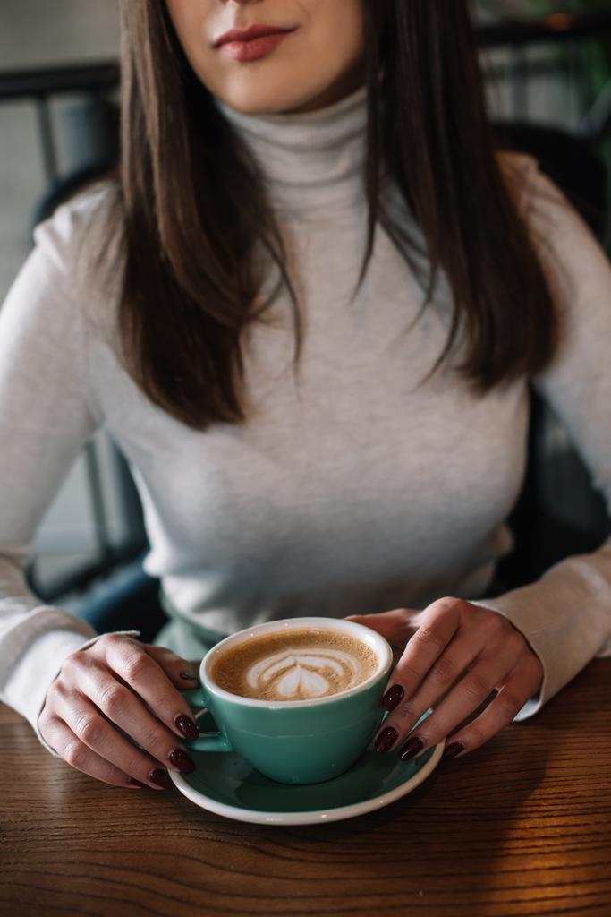 cropped view of young woman sitting at wooden table with cappuccino on balcony in coffee shop - Photo, Image