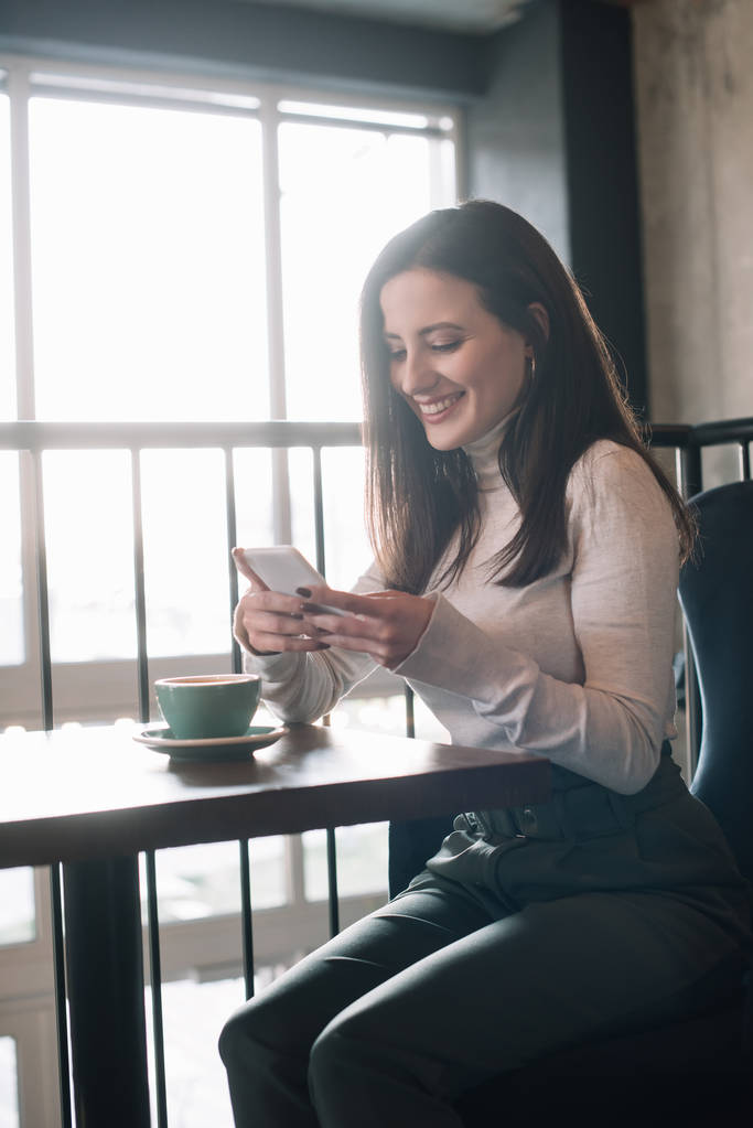 Gülümseyen genç bir kadın kapuçinolu ahşap masada oturuyor ve kahve dükkanının balkonunda akıllı telefon kullanıyor. - Fotoğraf, Görsel