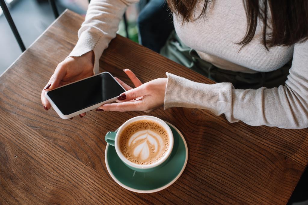 bijgesneden zicht op jonge vrouw zitten aan houten tafel met cappuccino en houden smartphone met leeg scherm op balkon in coffeeshop - Foto, afbeelding