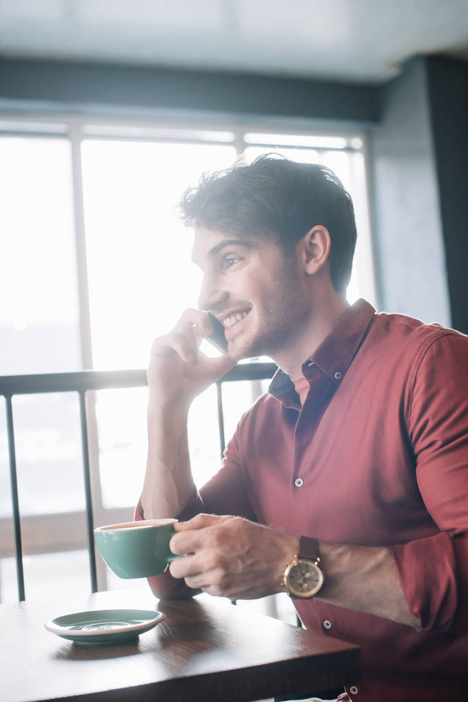 mosolygós fiatalember ül fa asztal cappuccino és beszél okostelefonon az erkélyen kávézóban - Fotó, kép