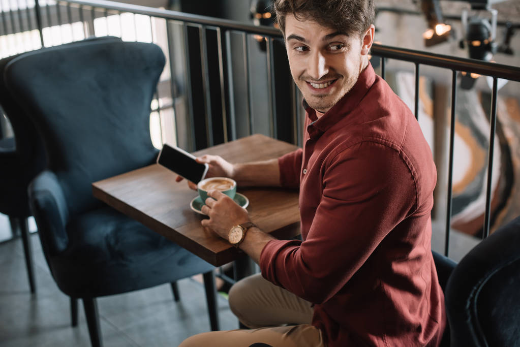 usměvavý mladý muž sedí u dřevěného stolu s cappuccino a drží smartphone na balkóně v kavárně - Fotografie, Obrázek