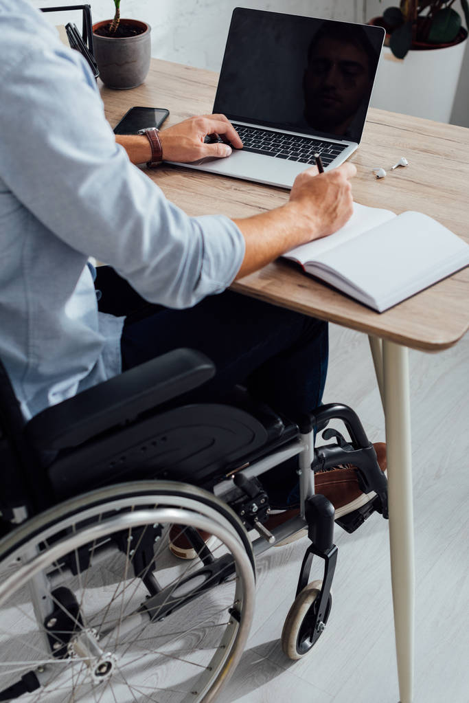 Cropped view of man in wheelchair writing in notebook and using laptop at desk - Photo, Image