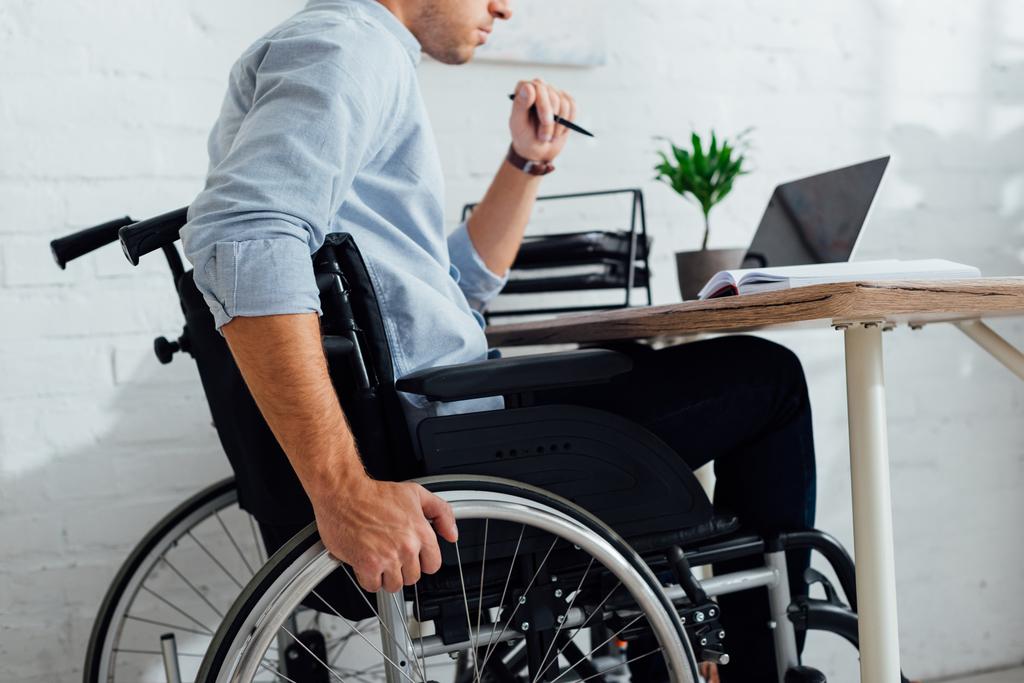 Vista recortada del hombre sentado en silla de ruedas por cuaderno en el lugar de trabajo
 - Foto, Imagen