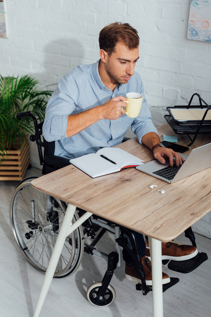 Άνδρας σε αναπηρική καρέκλα χρησιμοποιώντας φορητό υπολογιστή και πίνοντας καφέ στο χώρο εργασίας - Φωτογραφία, εικόνα