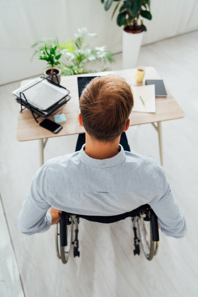 Zadní pohled na muže na invalidním vozíku sedí u stolu s notebookem a papíry - Fotografie, Obrázek