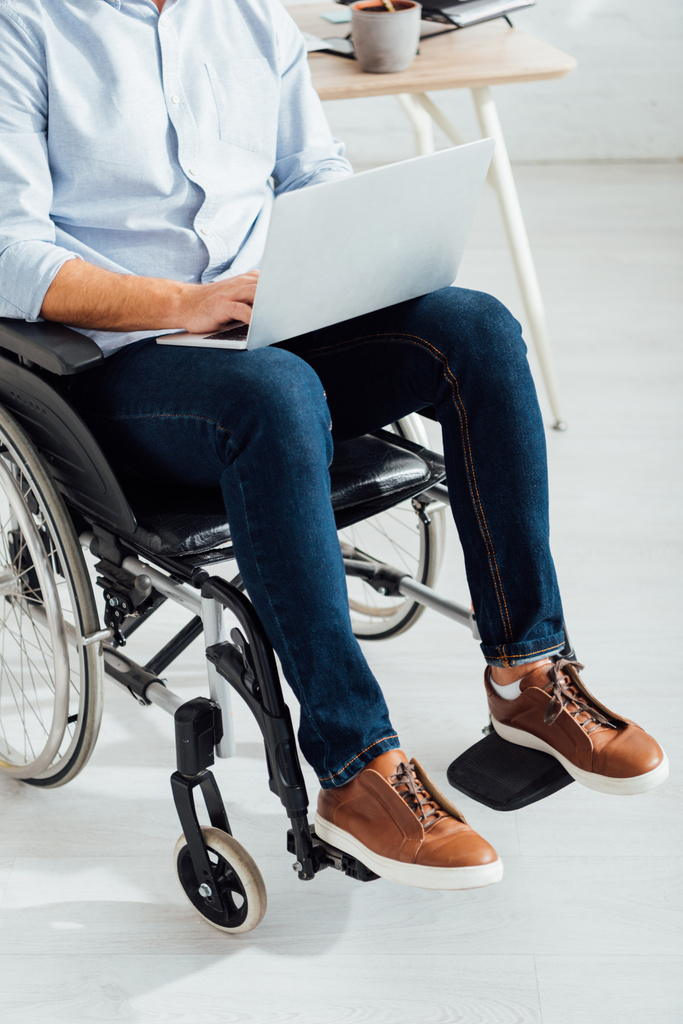 Обрізаний вид людини, що сидить у інвалідному кріслі та використовує ноутбук
 - Фото, зображення