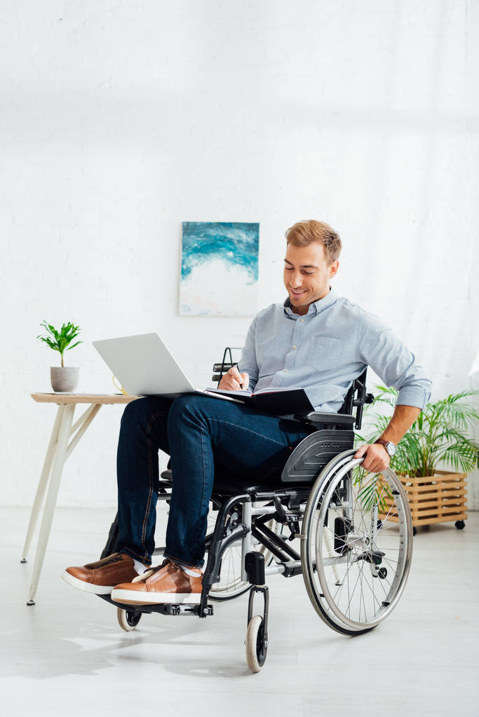 Glimlachende man in een rolstoel die schrijft in een notitieboekje en laptop vasthoudt in de woonkamer - Foto, afbeelding