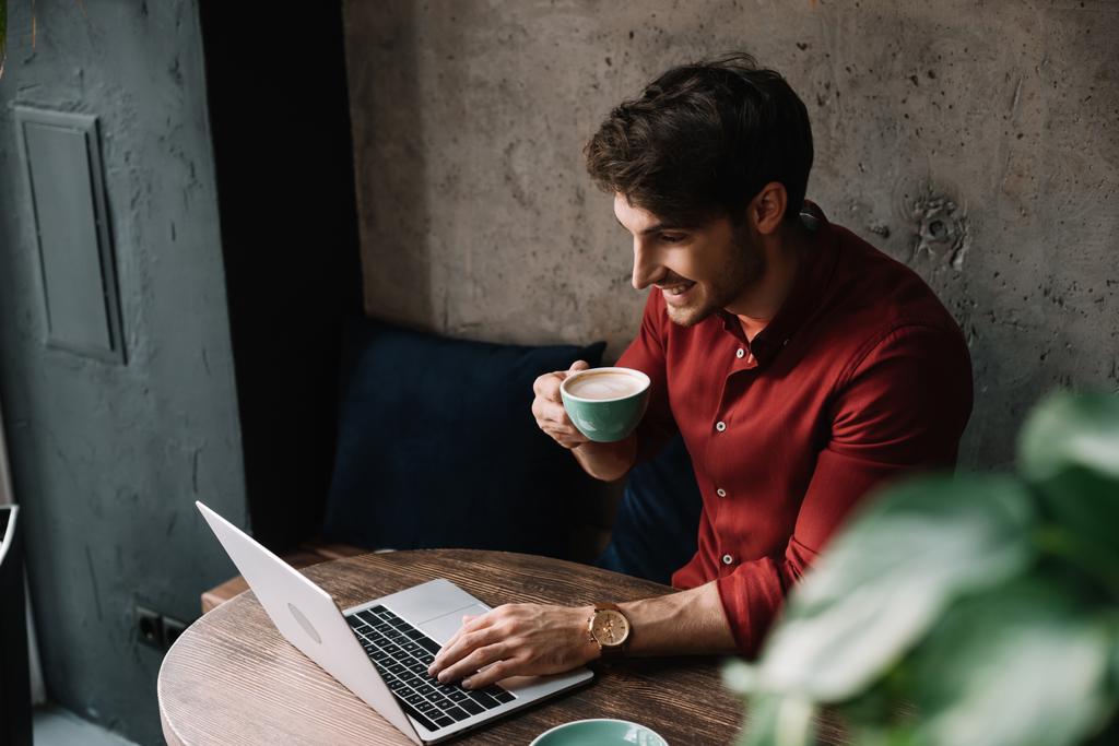 glimlachende jonge freelancer werkt op laptop en het drinken van koffie in de coffeeshop - Foto, afbeelding