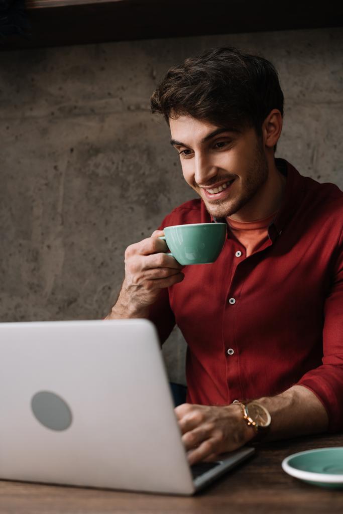 コーヒーショップでノートパソコンやコーヒーを飲みながら働く若いフリーランサーの笑顔 - 写真・画像