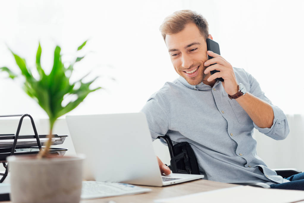 Hymyilevä freelancer puhuu älypuhelimella ja käyttää kannettavaa tietokonetta pöydällä
 - Valokuva, kuva