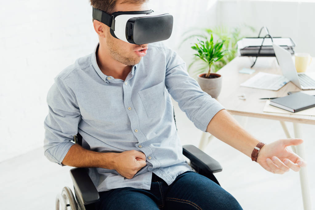 Uomo eccitato in sedia a rotelle utilizzando cuffie realtà virtuale dal posto di lavoro
 - Foto, immagini